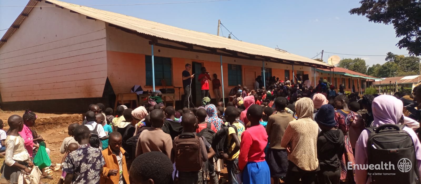 A crowd of children and adults gathered in front of a school building, facing a speaker.